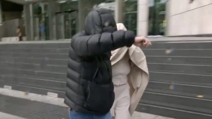 Two pedestrians on a city sidewalk; one wears a dark puffer jacket with the hood up, the other wears a beige coat and covers part of their face.