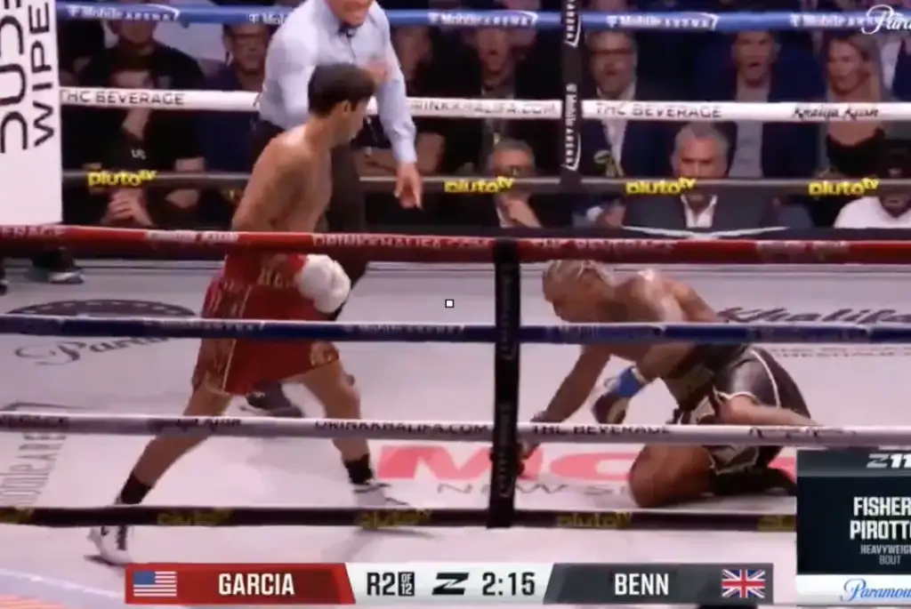 Boxer Garcia stands in red trunks facing Benn, who is kneeling on the canvas in a boxing ring during the second round edge of the ring.