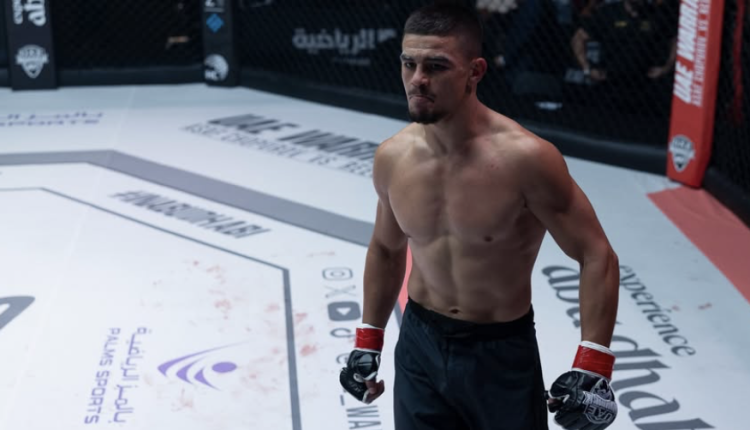 Male MMA fighter in a cage, shirtless, wearing red gloves and black shorts, looking forward.