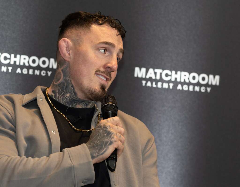Man with tattoos and a beige jacket speaks into a handheld microphone against a dark backdrop with Matchroom branding.