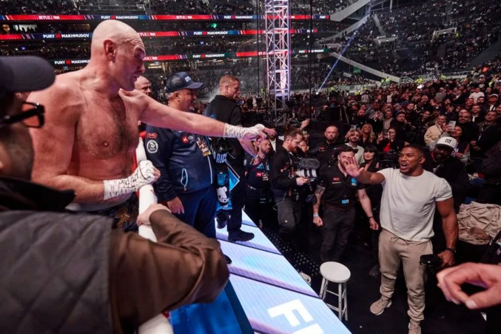 Bald boxer reaches to high-five a spectator in a white shirt at ringside as a packed arena cheers.