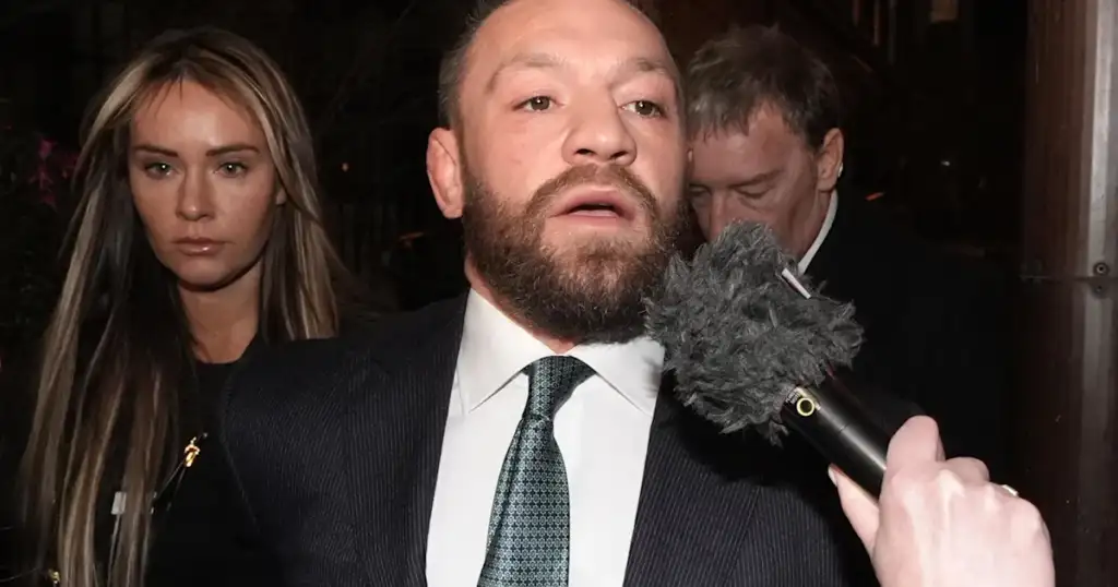 Bearded man in a suit being interviewed by a handheld microphone; a woman stands behind him watching.