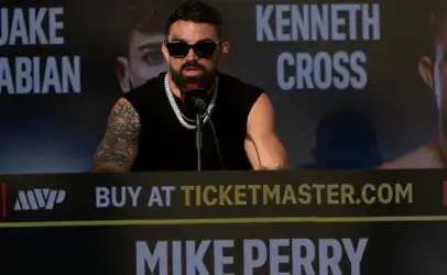 Boxer Mike Perry at a press conference, wearing sunglasses and a black sleeveless shirt, speaking into a microphone at a promotional backdrop.