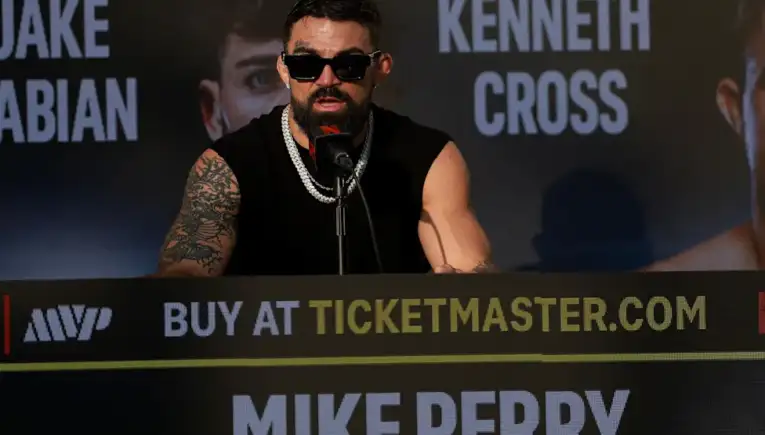 Boxer Mike Perry at a press conference, wearing sunglasses and a black sleeveless shirt, speaking into a microphone at a promotional backdrop.