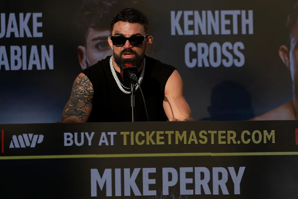 Boxer Mike Perry at a press conference, wearing sunglasses and a black sleeveless shirt, speaking into a microphone at a promotional backdrop.