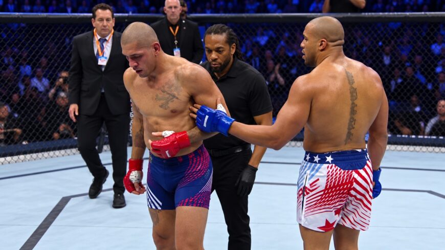 Two shirtless mixed martial artists in a cage after a bout, one with red gloves and blue shorts, the other with blue gloves and USA-themed shorts, a referee nearby.