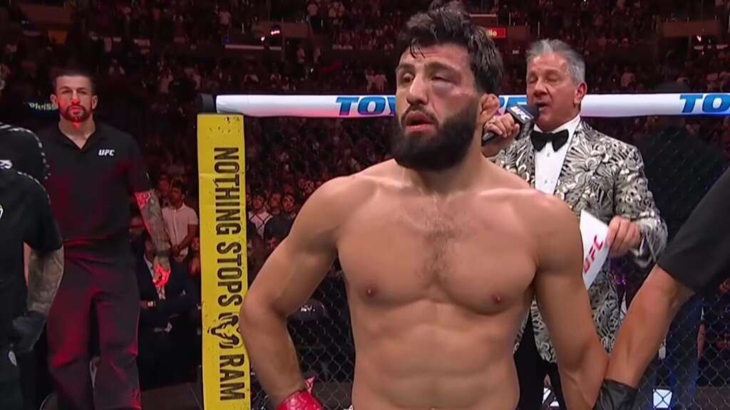 Shirtless male mixed martial artist in the octagon holds hands with a teammate while a presenter speaks in a patterned suit nearby.