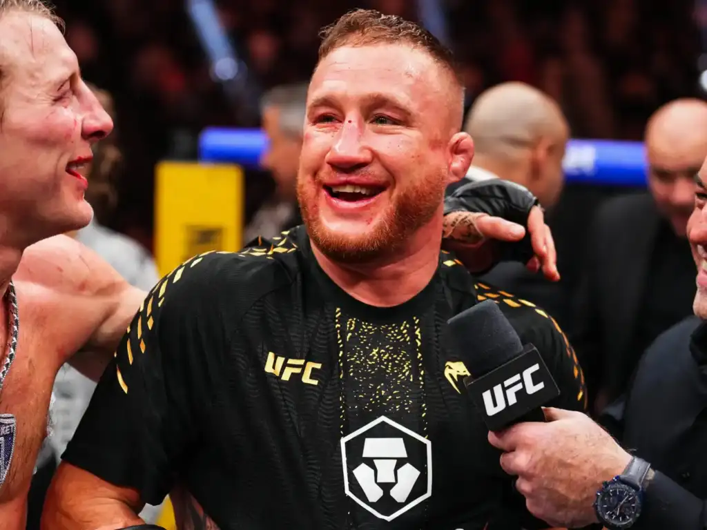 UFC fighter in a black and gold shirt smiles during a post-fight interview as a reporter holds a UFC microphone nearby.