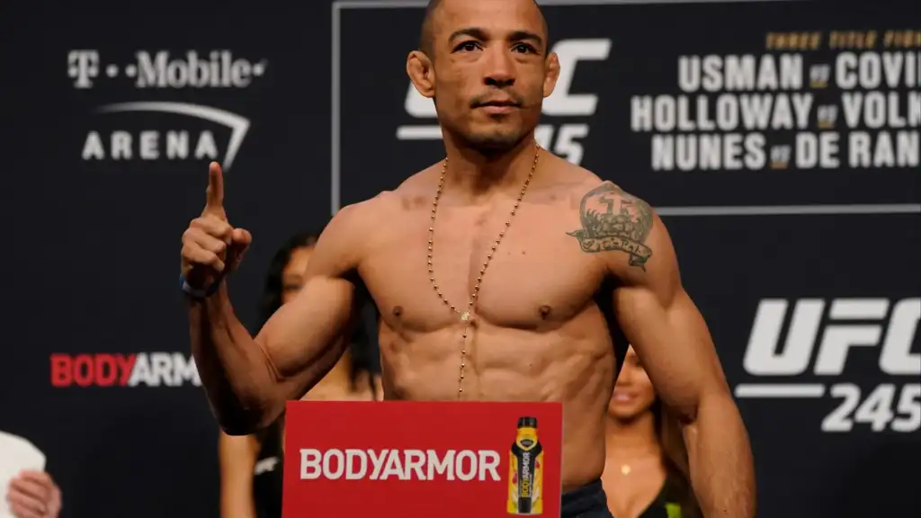Physically fit man on a weigh-in stage pointing upward, a BodyArmor sign visible in front with a bottle image.