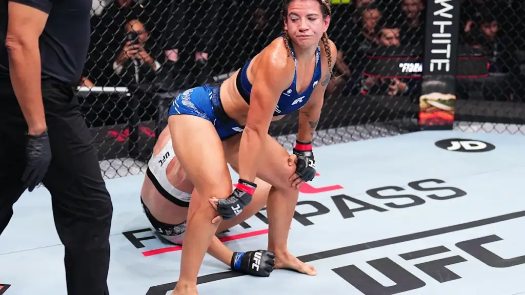 Female UFC fighter in blue shorts stands over a downed opponent inside the cage as a referee watches nearby.