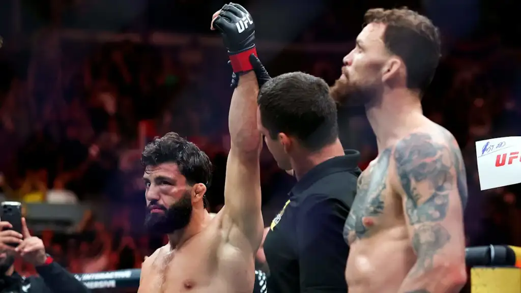 MMA fighter with beard raises his gloved hand in victory as a referee stands between him and a shirtless opponent, crowd in the arena in the background.