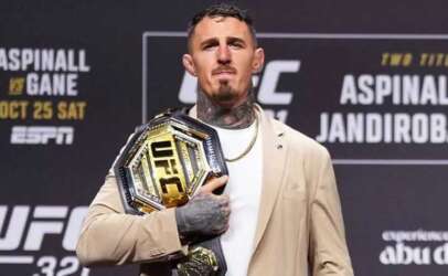 Male fighter standing on stage, holding the UFC championship belt over his chest, wearing a beige blazer.