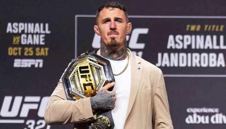 Male fighter standing on stage, holding the UFC championship belt over his chest, wearing a beige blazer.
