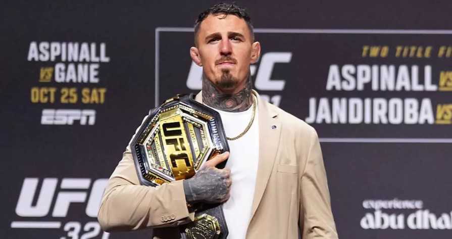 Male fighter standing on stage, holding the UFC championship belt over his chest, wearing a beige blazer.