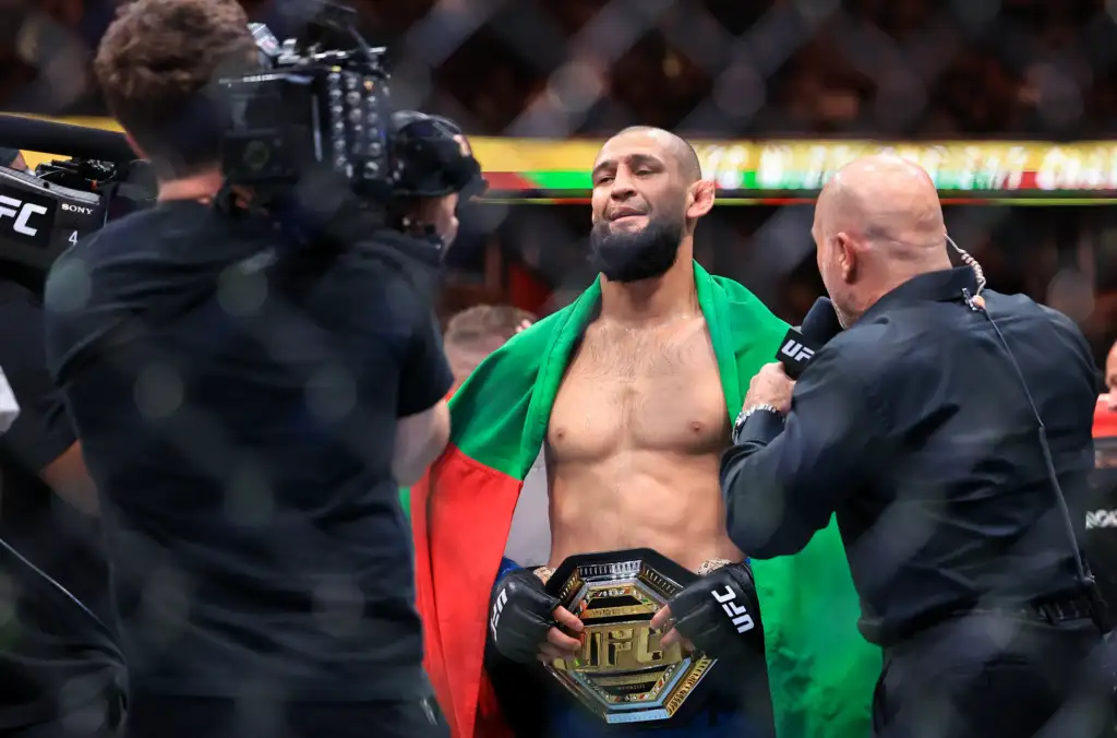 UFC fighter with a championship belt around his waist, draped in a green and red flag as cameras film him during an interview.