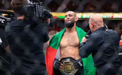 UFC fighter with a championship belt around his waist, draped in a green and red flag as cameras film him during an interview.