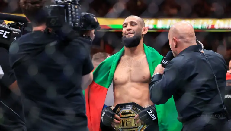UFC fighter with a championship belt around his waist, draped in a green and red flag as cameras film him during an interview.