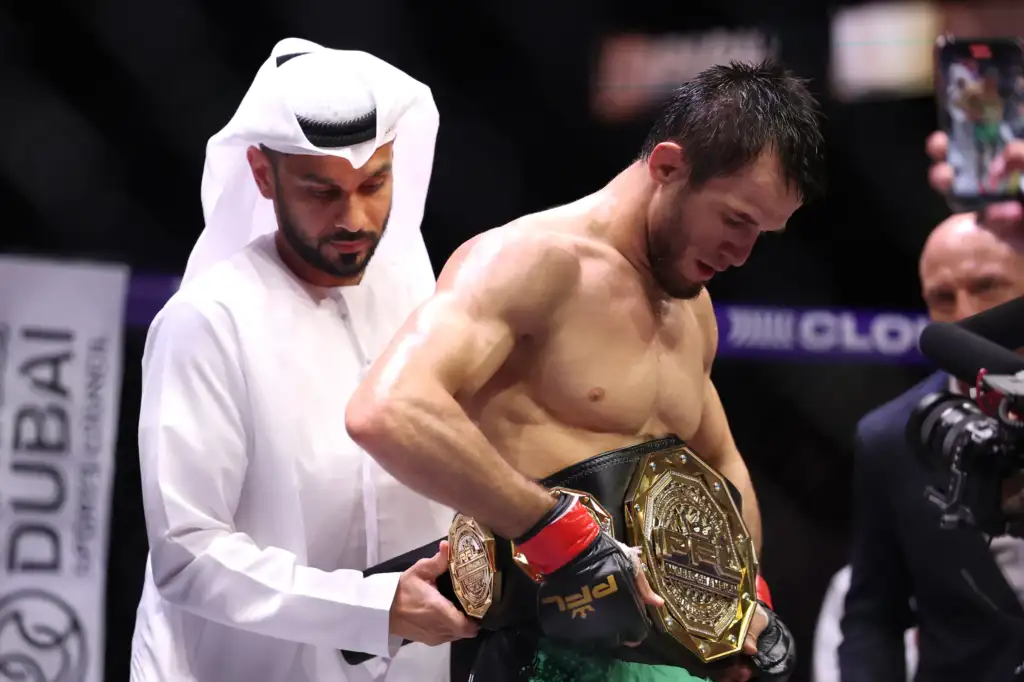 Shirtless mixed martial artist with multiple gold championship belts being adjusted by a man in white traditional clothing in a ring setting.