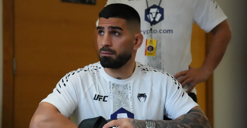 Man with short dark hair and beard wearing a white UFC shirt, looking to the left, arms resting on a surface; a person in a white shirt stands behind.