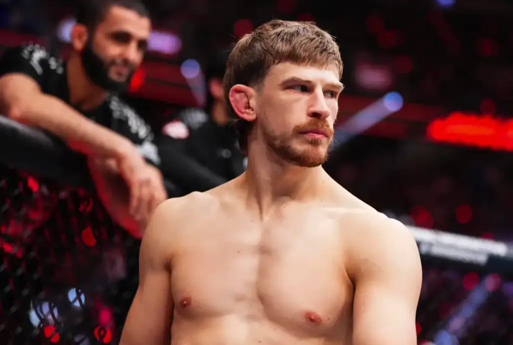 Shirtless male mixed martial artist standing by the cage, looking to the side with a focused expression; blurred spectators in the background.