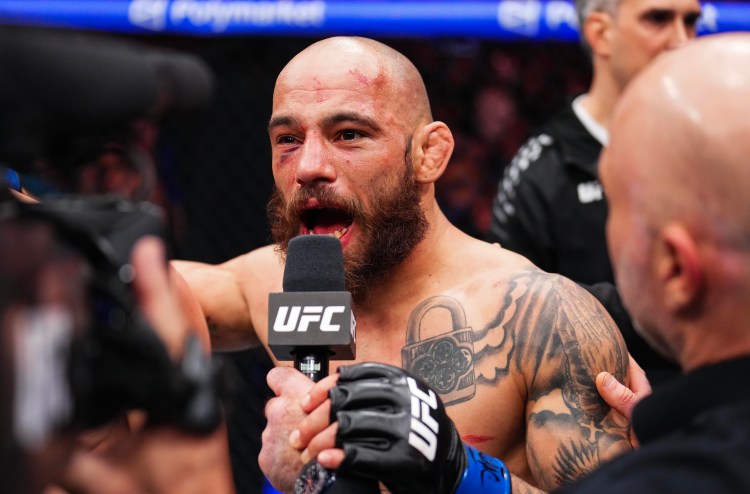 UFC fighter with a shaved head and beard speaks into a UFC microphone during an interview after a fight, his tattoos visible and blood on his face.