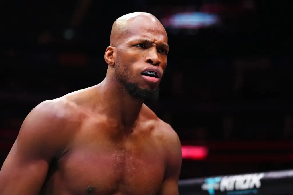 Close-up of a shirtless mixed martial artist wearing a UFC-branded mouthguard, focused in a dim arena.