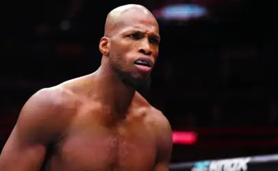Close-up of a shirtless mixed martial artist wearing a UFC-branded mouthguard, focused in a dim arena.