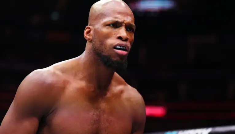 Close-up of a shirtless mixed martial artist wearing a UFC-branded mouthguard, focused in a dim arena.