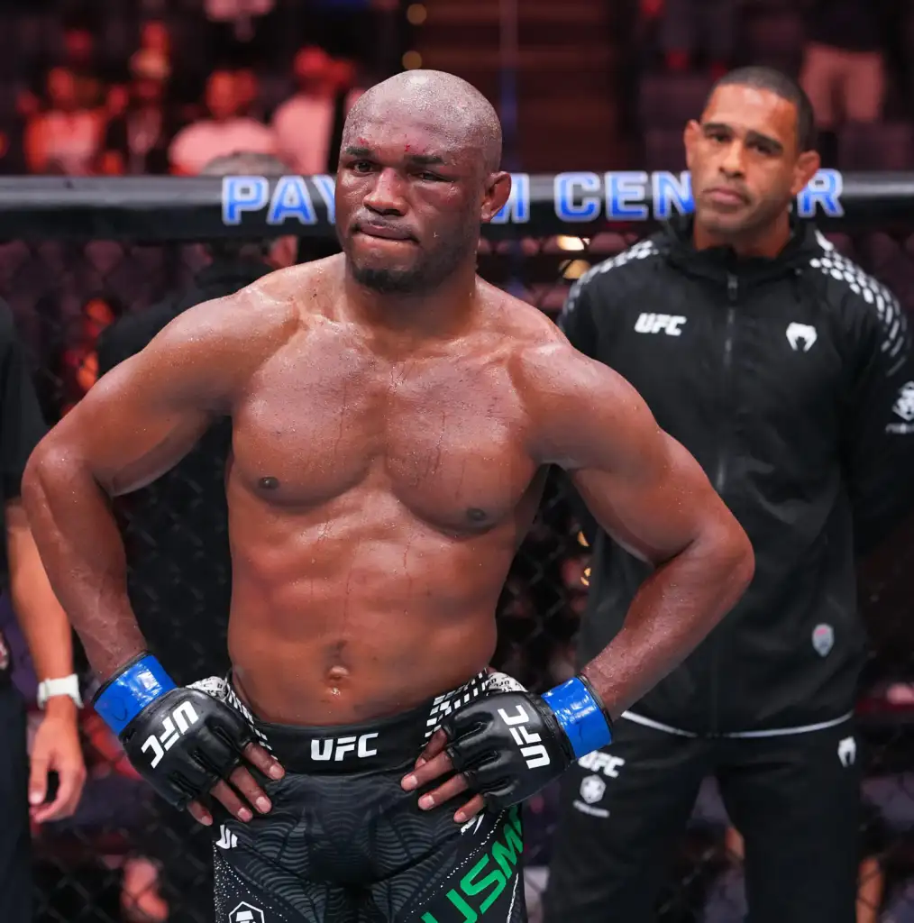 gettyimages-2286700935 - AfroCombat A shirtless UFC fighter stands in the octagon with hands on hips, wearing black gloves and shorts, a focused expression after a bout or weigh-in.