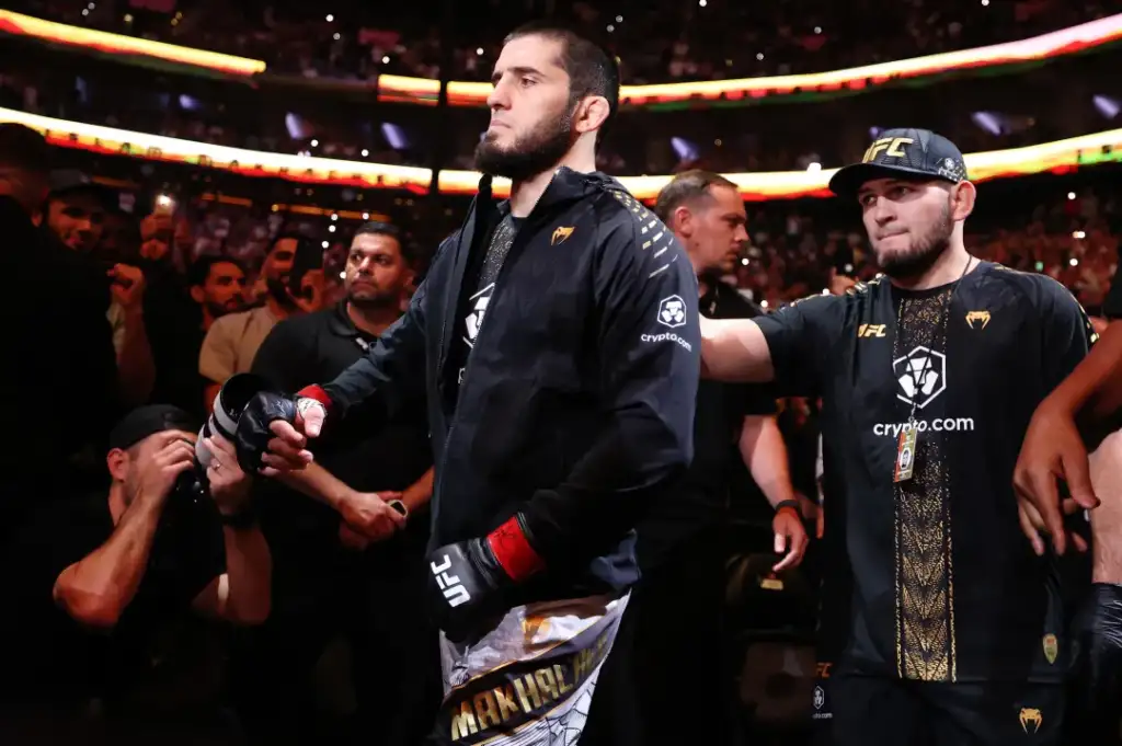 Two UFC fighters walk to the arena with a cheering crowd around them, one in a black jacket and red gloves flanked by teammates and photographers.