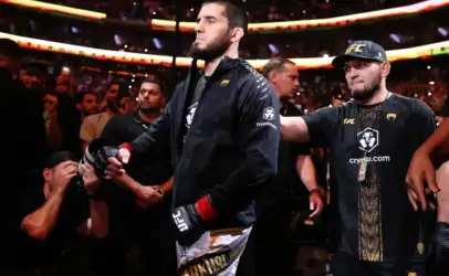 Two UFC fighters walk to the arena with a cheering crowd around them, one in a black jacket and red gloves flanked by teammates and photographers.