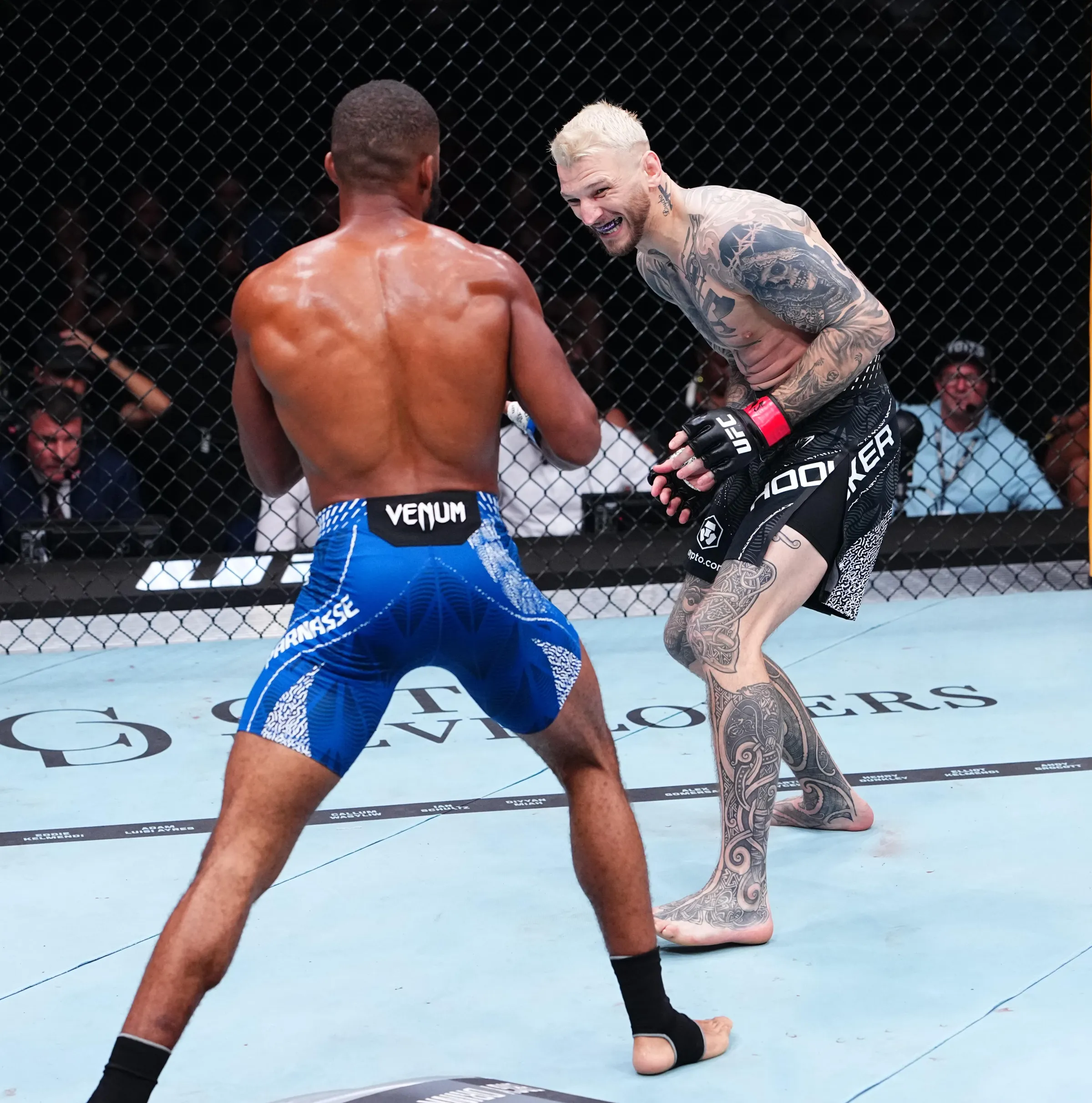 Two shirtless mixed martial artists face off inside a chain‑link cage, one with blue shorts and the other heavily tattooed.