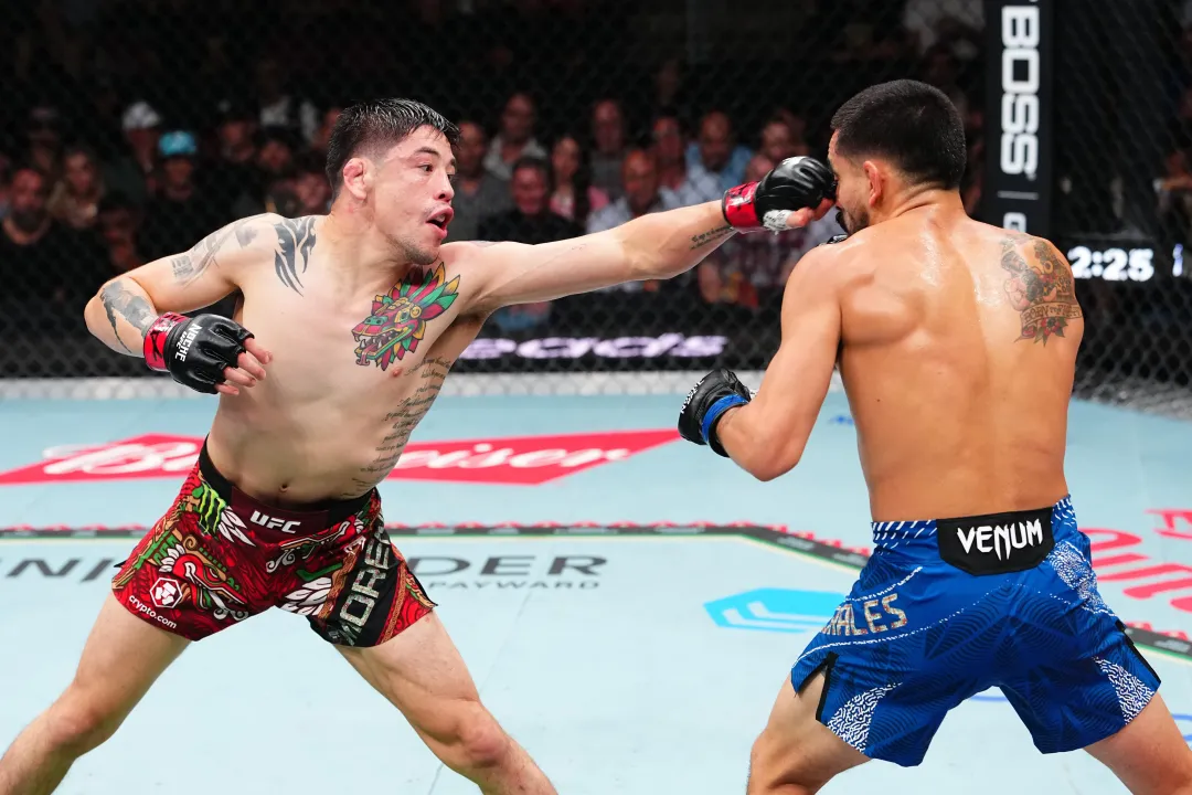 Two mixed martial artists exchange a punch in a UFC octagon, one in red shorts extending a left hand toward the opponent in blue shorts.
