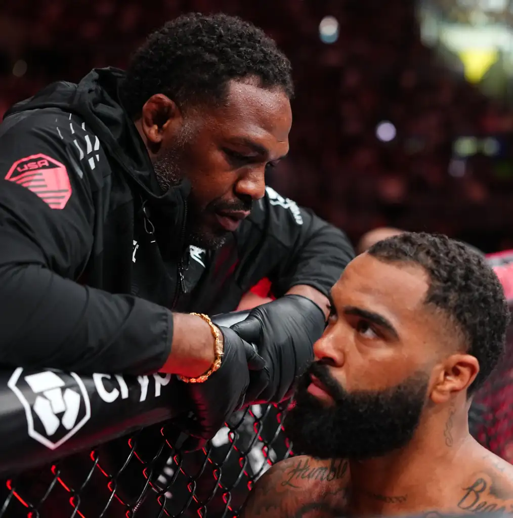 Man in black jacket leaning on a chain-link fence at an MMA event, with another man's head in foreground inside a cage.