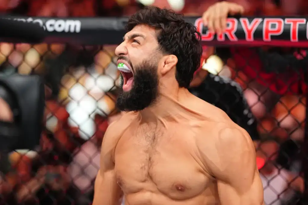 Muscular MMA fighter screams with mouthguard in a cage, fists clenched, behind a chain-link fence.