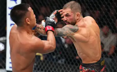 Two shirtless UFC fighters inside a cage trade punches, one delivering a right hand toward the other's head with tattooed arms visible.