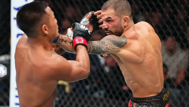 Two shirtless UFC fighters inside a cage trade punches, one delivering a right hand toward the other's head with tattooed arms visible.