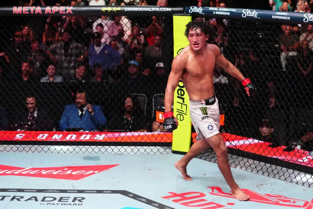 Male UFC fighter, shirtless with red gloves, walking inside the octagon near the cage as the crowd watches.