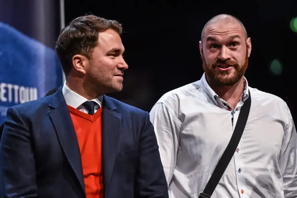Two men stand on a stage, one in a navy blazer with an orange sweater, the other in a white shirt with a black crossbody strap, chatting politely.
