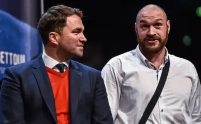 Two men stand on a stage, one in a navy blazer with an orange sweater, the other in a white shirt with a black crossbody strap, chatting politely.