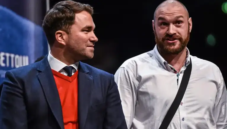 Two men stand on a stage, one in a navy blazer with an orange sweater, the other in a white shirt with a black crossbody strap, chatting politely.