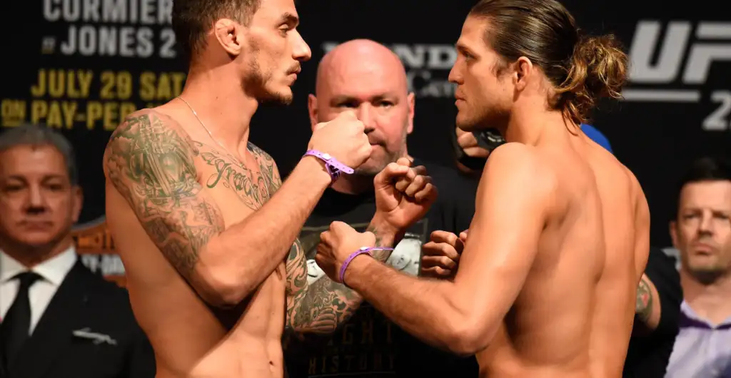 Two shirtless fighters face off in a weigh-in, their fists inches apart as an official and onlookers watch in the background.