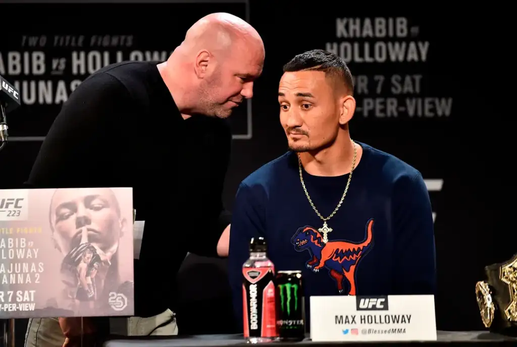 A bald man in a black shirt leans toward UFC fighter Max Holloway, who sits at a table during a press event wearing a navy shirt with a dinosaur graphic and a gold chain. On the table are a UFC poster, beverage cans, and a name plate reading 'MAX HOLLOWAY'.