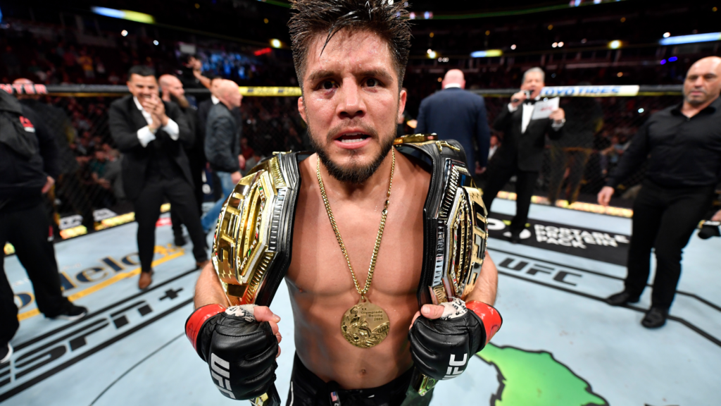 UFC fighter with multiple gold championship belts draped over shoulders, standing in the octagon with a determined expression.