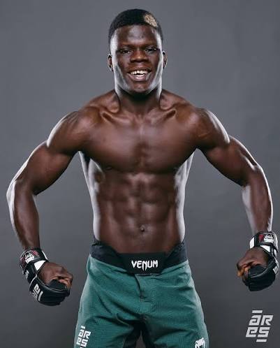 Muscular Black male athlete in green shorts wearing MMA gloves, standing with fists raised and smiling in a gray studio backdrop.