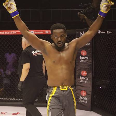Shirtless male mixed martial artist raises arms in victory inside a cage, wearing gray shorts with yellow trim and purple gloves.