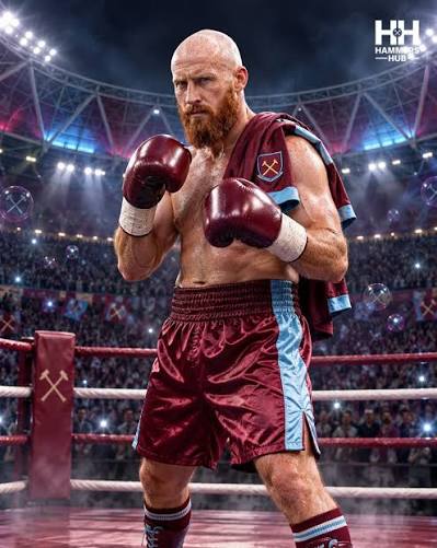 Boxer with a bald head and red beard stands in a boxing ring, gloves up, wearing maroon trunks with a light blue stripe and a towel over his shoulder.