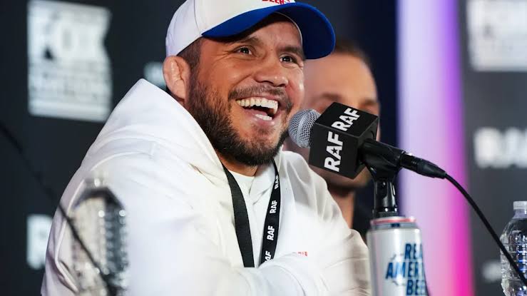 Smiling man in a white hoodie and blue-white cap speaks into a microphone at a panel.
