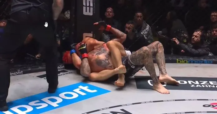 Two mixed martial artists on the mat trading ground-and-pound as a referee watches from the side inside the cage.