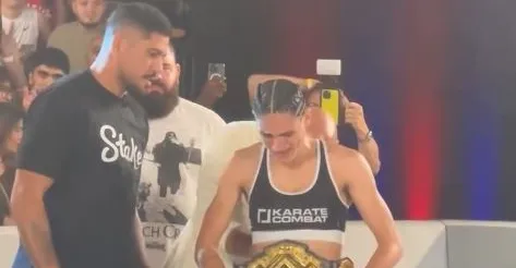 Female karate competitor wearing a black-and-white top and a championship belt, smiling in the ring as a man in a black shirt stands beside her; crowd in the background filming with phones.